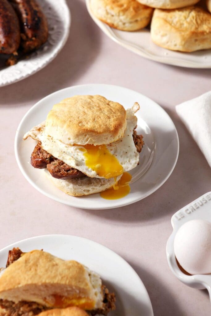 Two breakfast sandwiches on biscuits with a fried egg on white plates