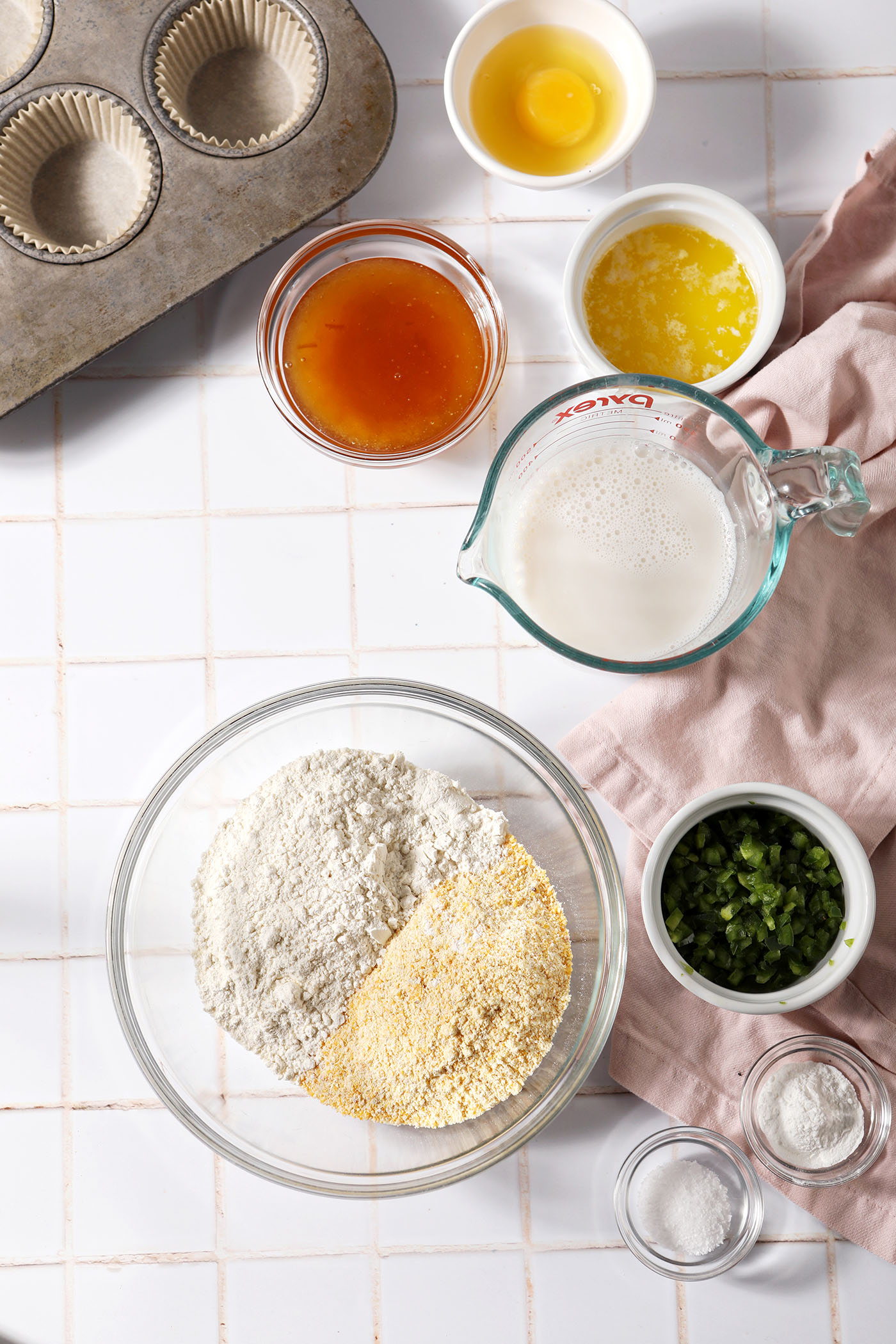 Ingredients to make cornbread muffins with honey and jalapeño on a white tiled surface in bowls next to a pink linen