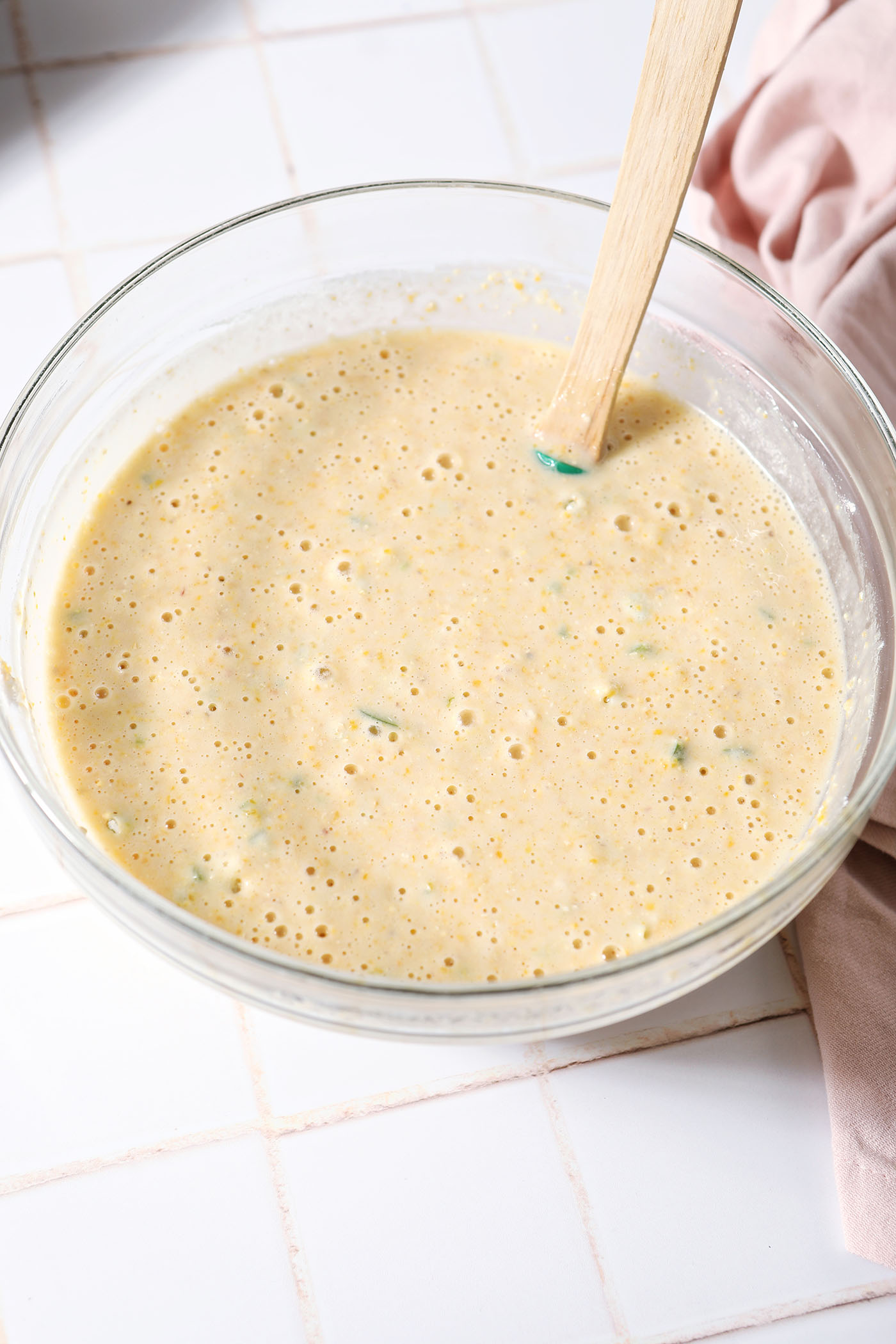 Mixed corn muffin batter in a bowl on a white tiled surface next to a pink linen