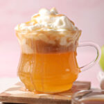 Close up of a spiked caramel apple cider topped with whipped cream on a wooden coaster on a white platter
