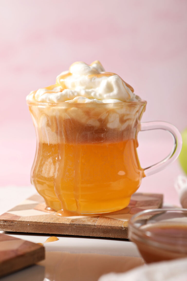 Close up of a spiked caramel apple cider topped with whipped cream on a wooden coaster on a white platter