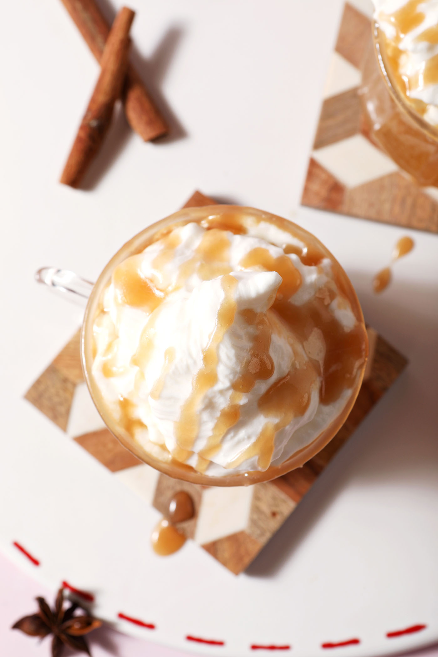 Overhead of a mug of spiked caramel apple cider topped with whipped cream on a wooden coaster on a white platter