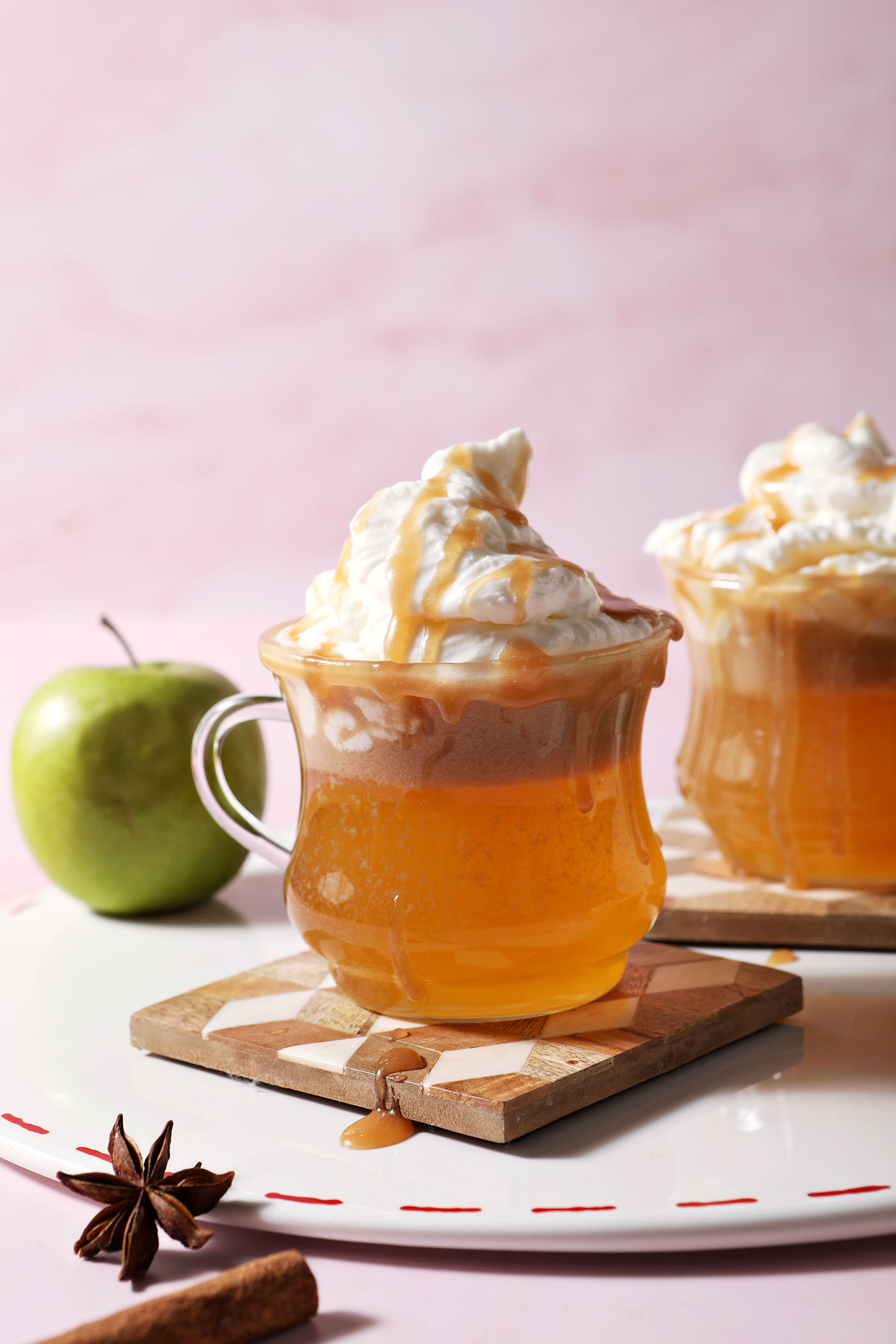 Two spiked caramel apple ciders topped with whipped cream and caramel drizzles in clear glass mugs on a white surface