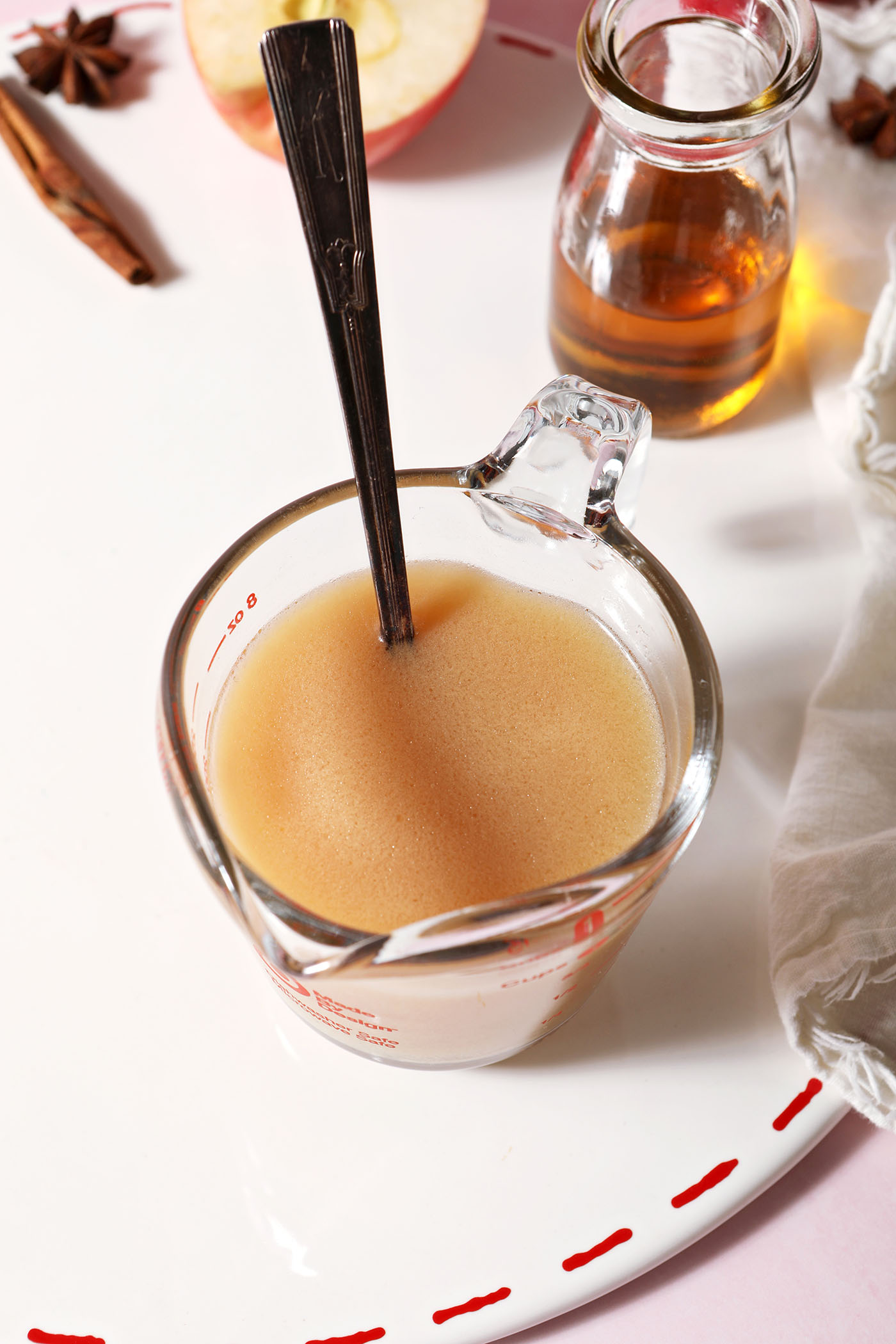Apple cider and caramel in a small measuring cup with a spoon on a white surface