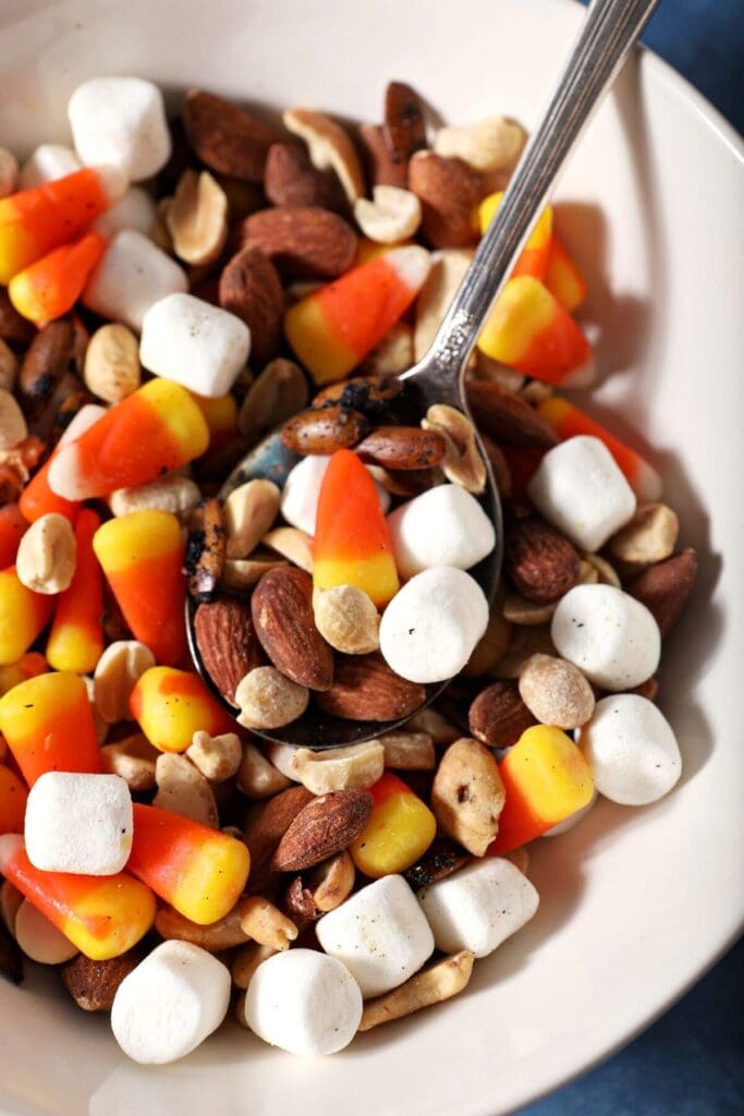 A spoon sits inside a bowl of nutty trail mix with candy corn and marshmallows