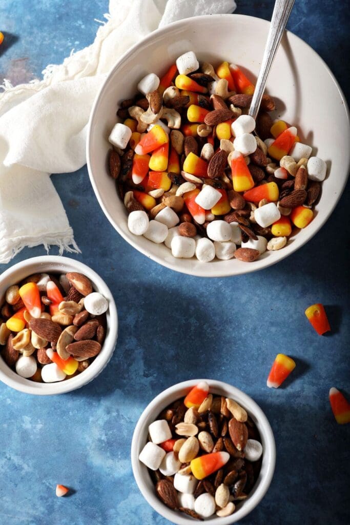 Two small bowls and one larger bowl of Halloween trail mix on a blue surface