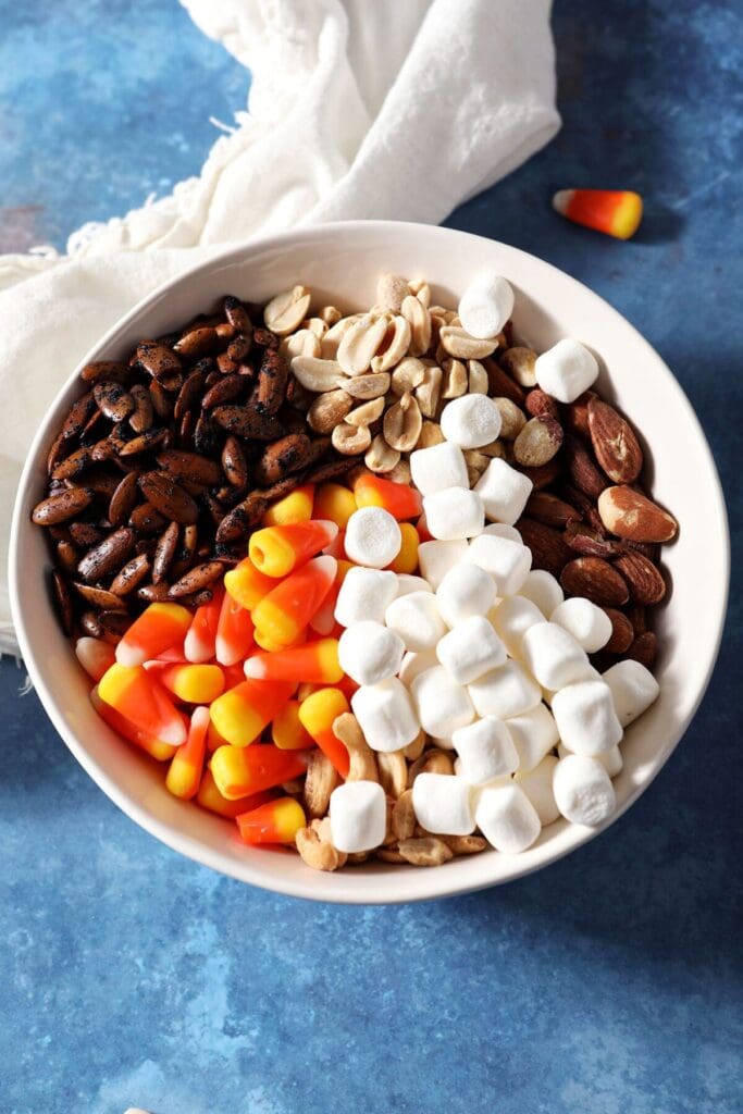 Ingredients stacked in a white bowl to make snack mix with candy corn and marshmallows