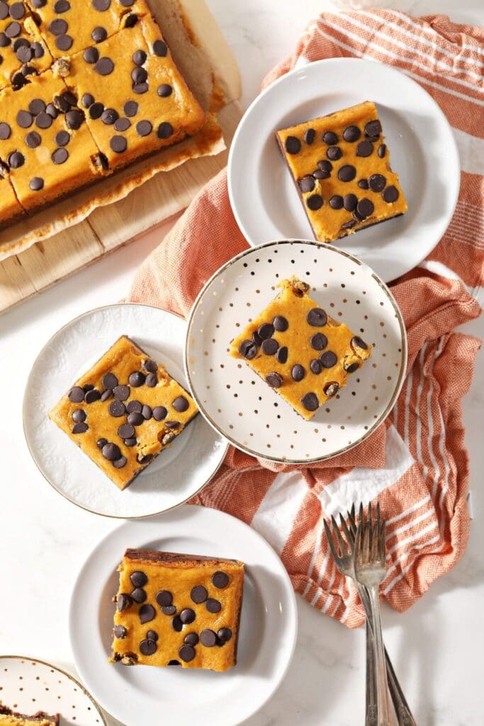 Five plates holding slices of Pumpkin Cheesecake Bars near a wooden platter of the rest of the brownie bars with a white and orange striped towel