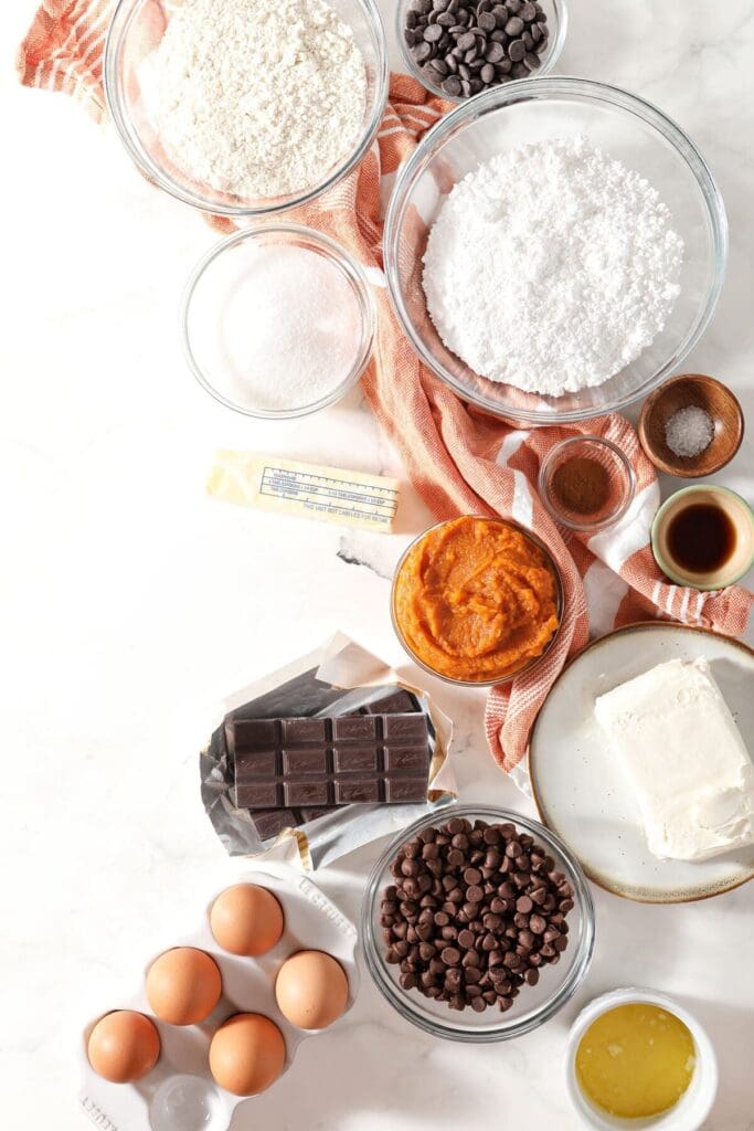 Ingredients to make Pumpkin Cheesecake Brownie Bars in bowls on a marble surface with a white and orange striped towel