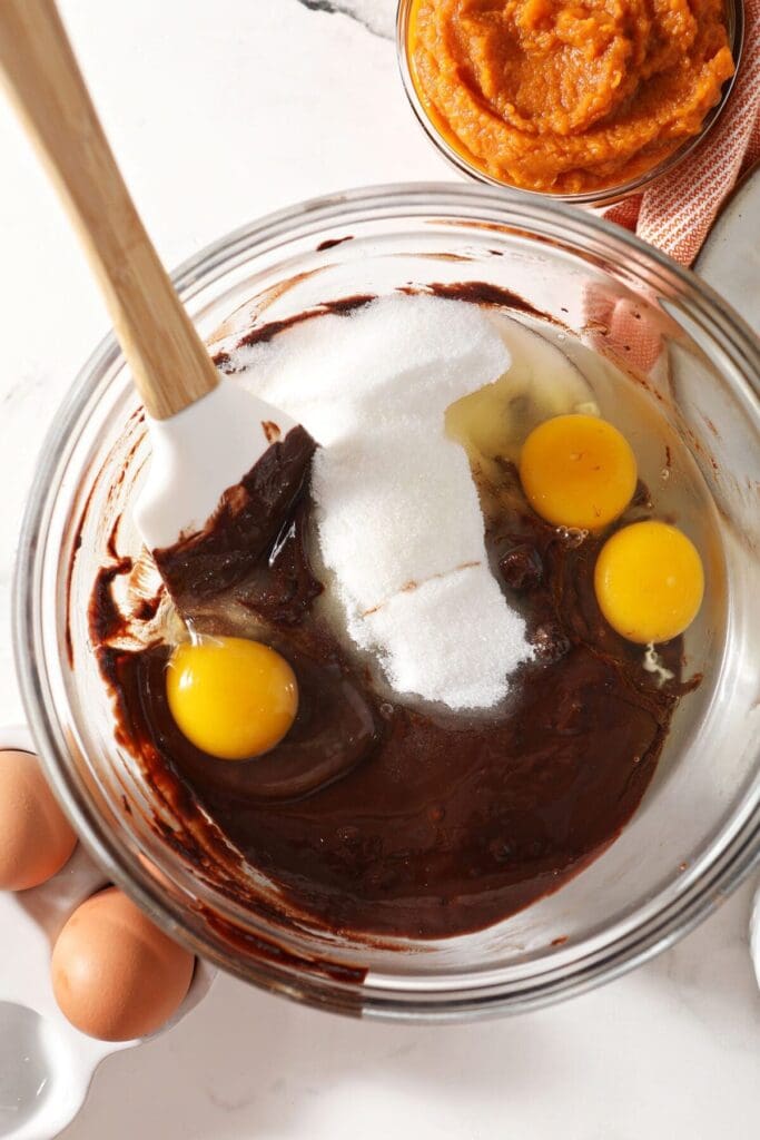 Sugar and eggs added to the chocolate mixture in a bowl with a rubber spatula