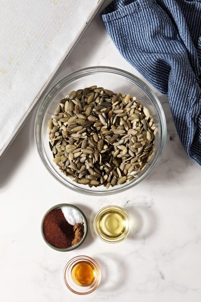 Ingredients to make roasted pumpkin seeds in bowls next to a parchment-lined sheet pan