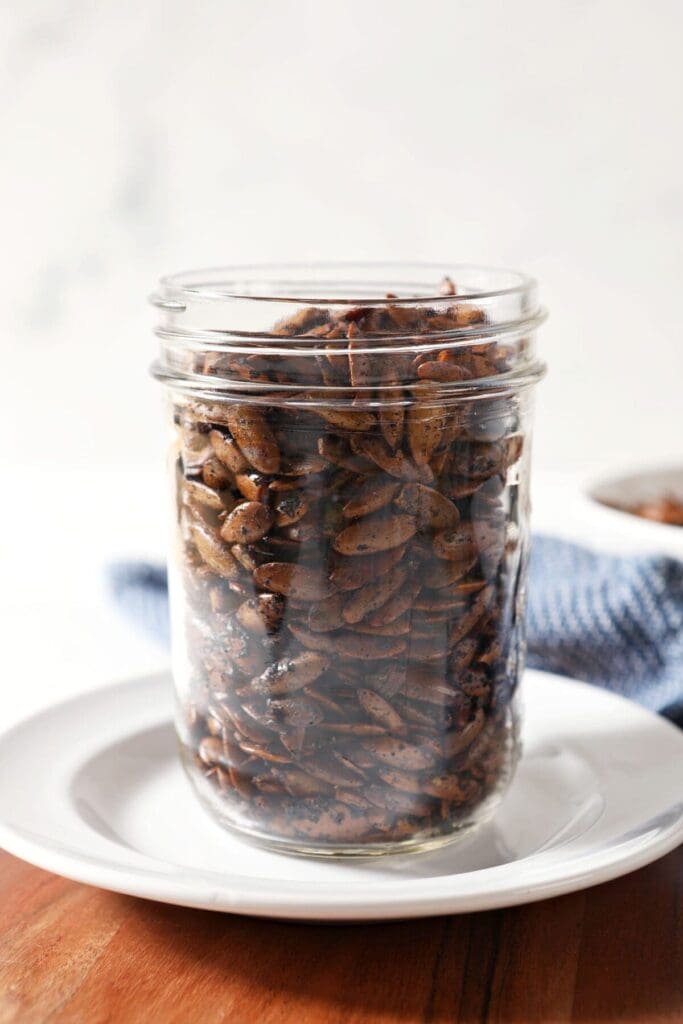 A mason jar of roasted pepitas on top of a white plate next to a blue towel