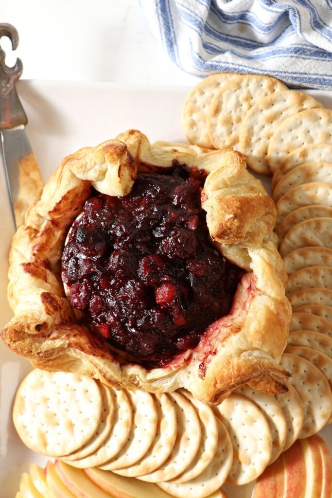 A cranberry Baked Brie wrapped in puff pastry surrounded by crackers and apple slices on a white platter