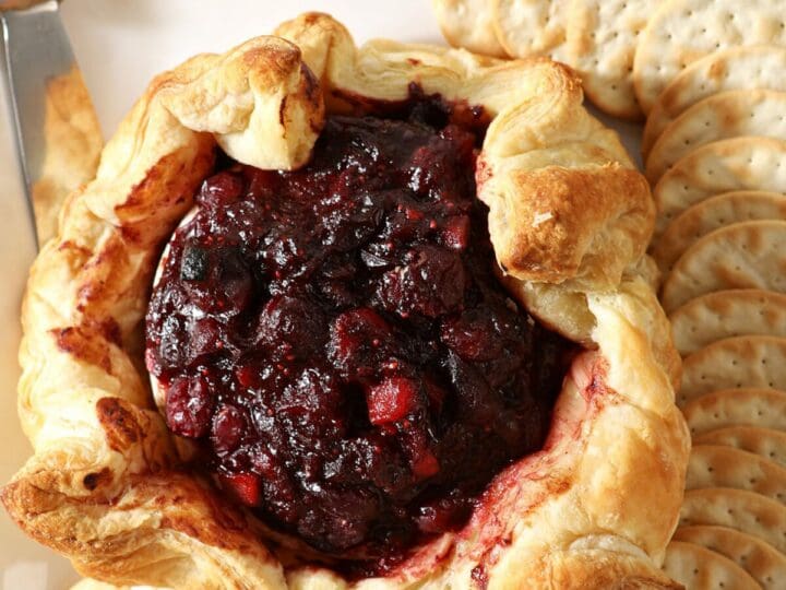 A cranberry Baked Brie wrapped in puff pastry surrounded by crackers and apple slices on a white platter
