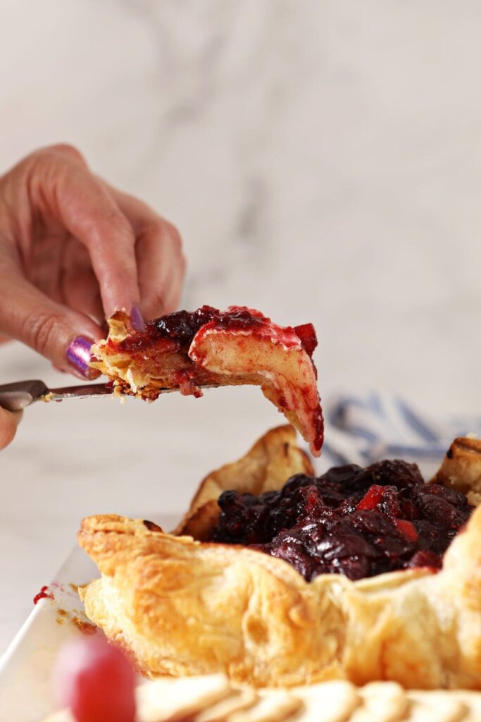 A hand lifts a slice of cranberry Baked Brie in puff pastry from a wheel of brie