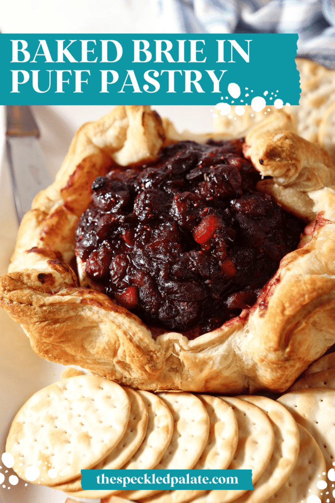 A cranberry Baked Brie wrapped in puff pastry surrounded by crackers and apple slices on a white platter with the text Baked Brie in puff pastry