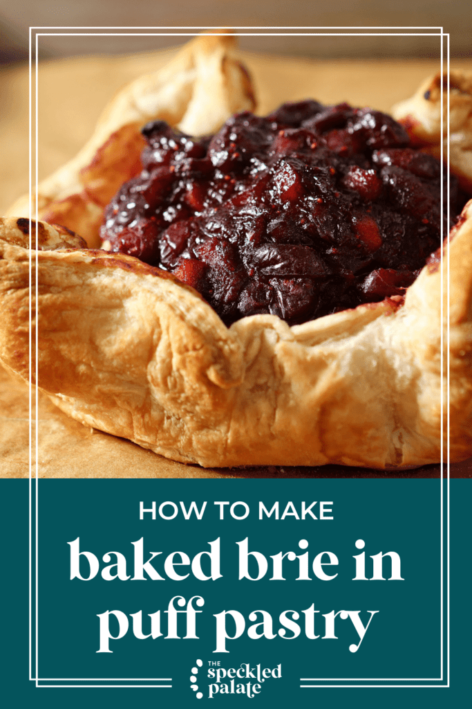 Baked Brie topped with cranberry chutney and wrapped in puff pastry sits on a parchment-lined sheet pan after baking with the text how to make Baked Brie in puff pastry