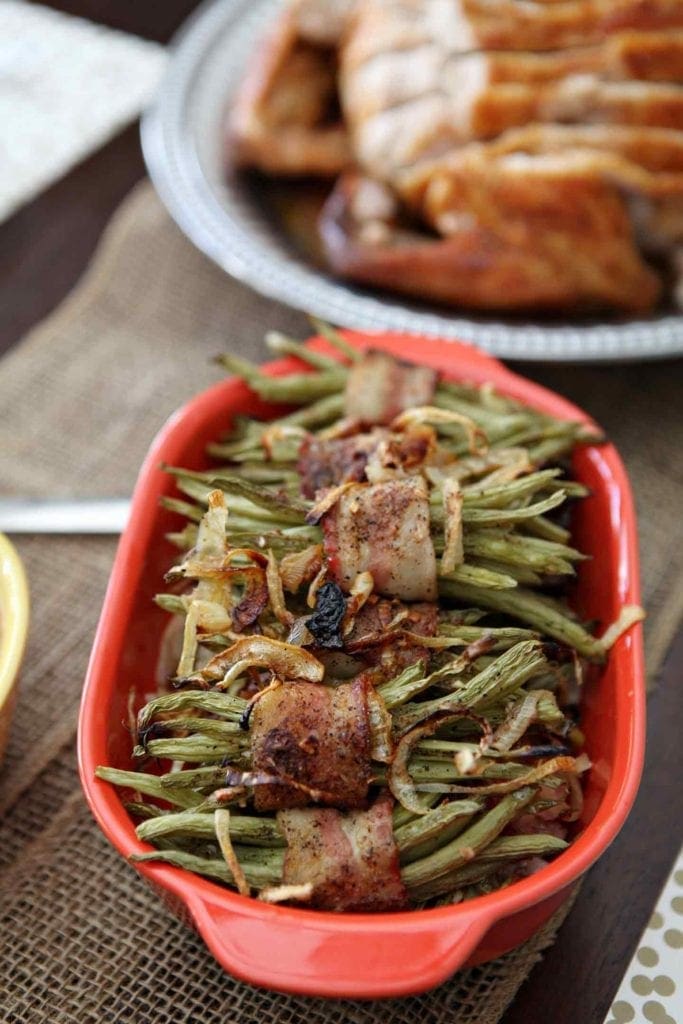 A red serving dish holds several Green Bean Bacon Bundles on a holiday table