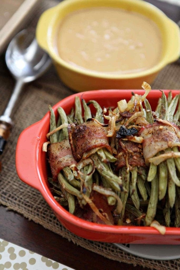 Several Green Bean Bacon Bundles sit in a red baking dish and are served alongside gravy at a holiday table