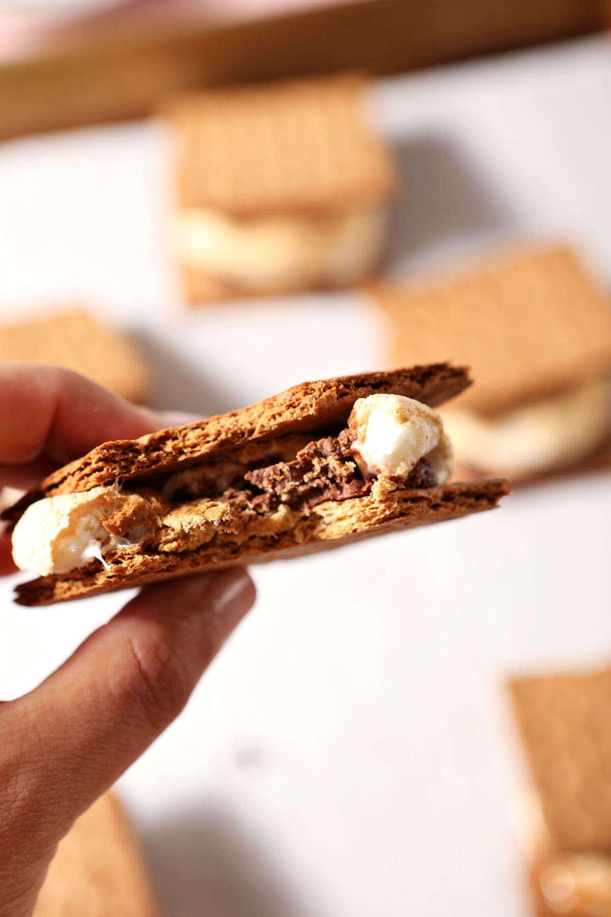A hand holds a bitten-into s’more above a sheet pan with more baked s’mores