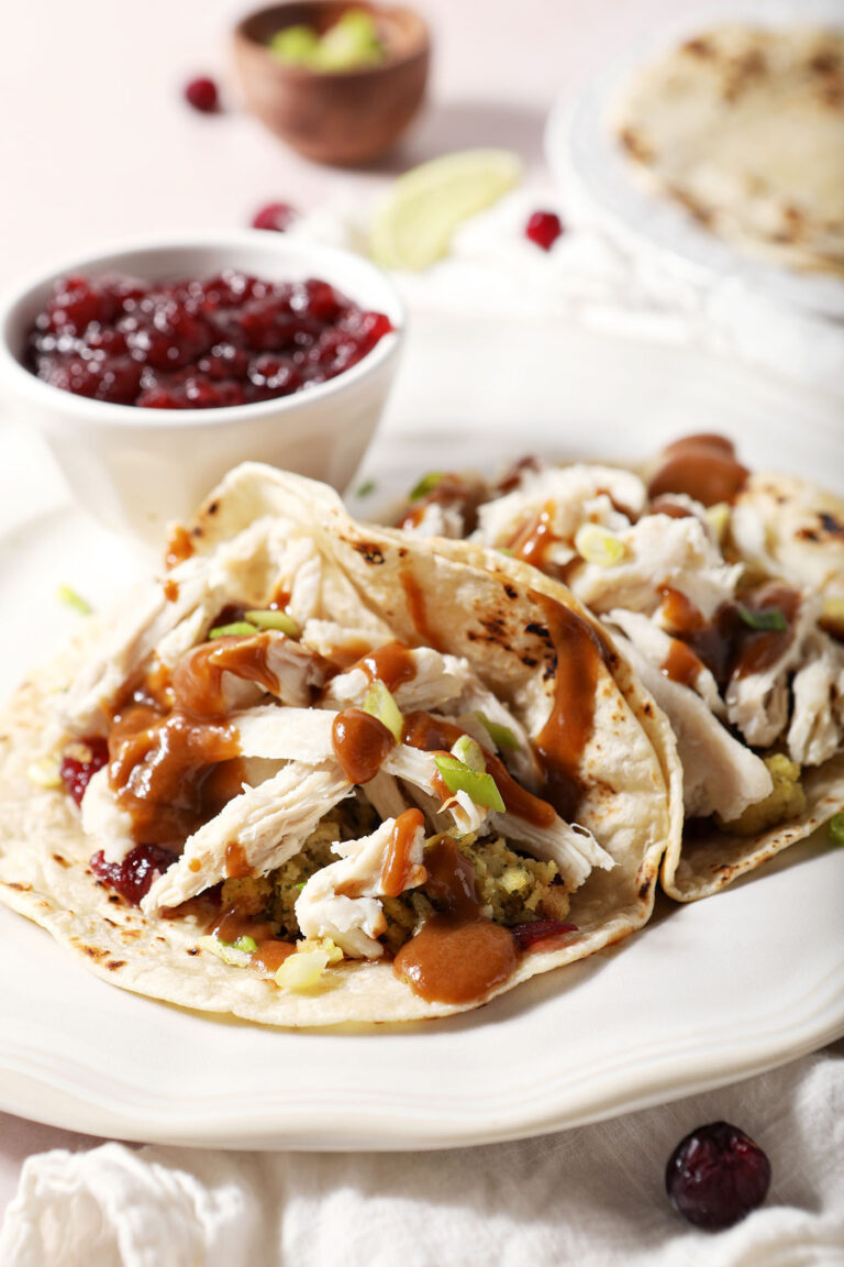 Side angle of two thanksgiving tacos topped with gravy, turkey, stuffing and cranberry sauce on a white plate next to a bowl of cranberry sauce