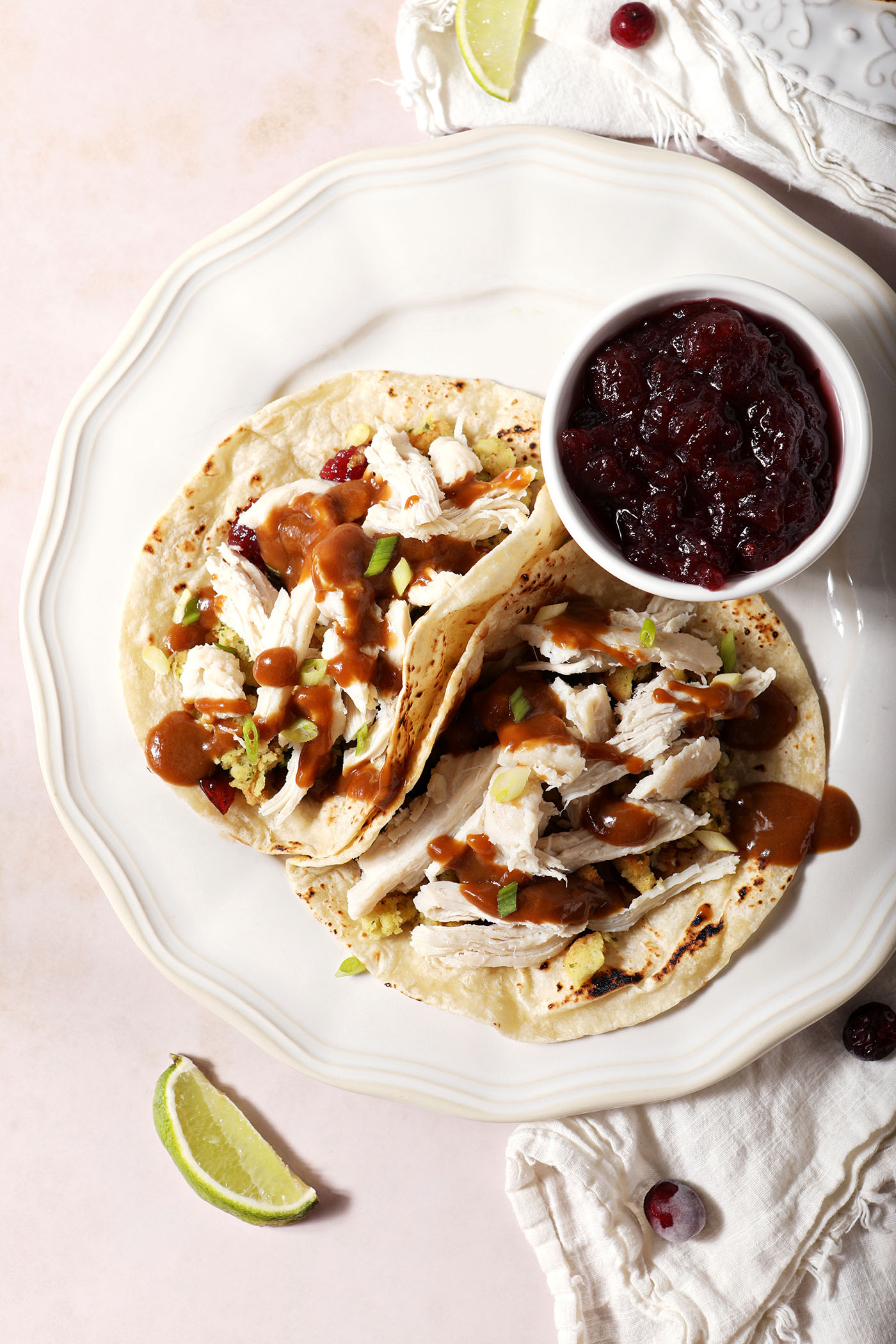 Overhead of two leftover tacos with turkey, gravy, stuffing and cranberry sauce on a white plate next to a bowl of cranberry sauce