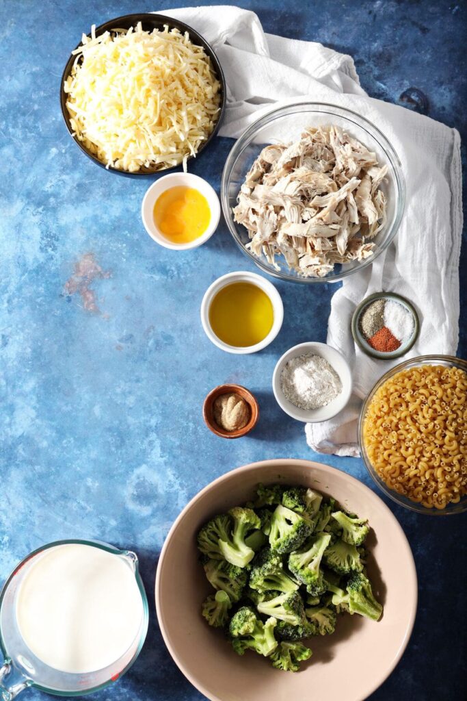 Ingredients to make homemade Mac ’n cheese with broccoli and chicken in bowls on a blue countertop