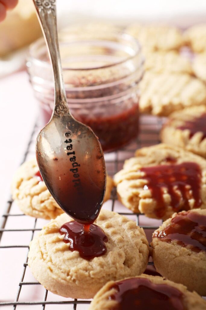 A spoon drizzles raspberry glaze onto peanut butter shortbread cookies on a cooling rack