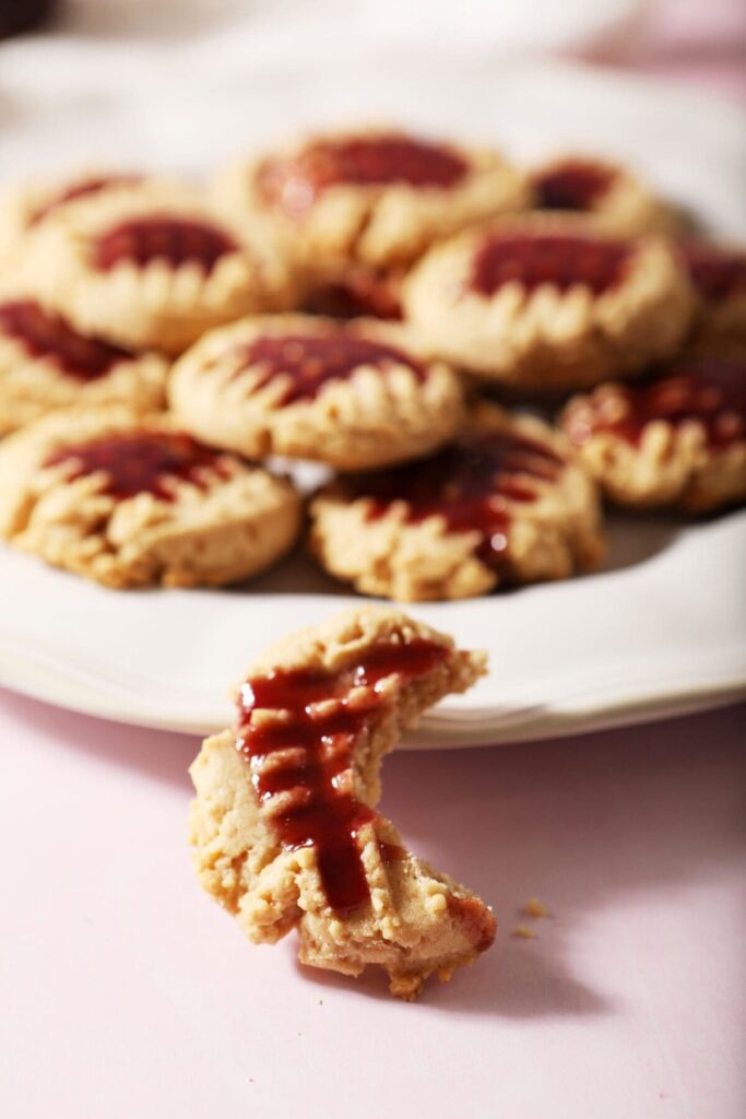 A bitten into Peanut Butter Shortbread Cookie glazed with raspberry leans against a plate with other cookies