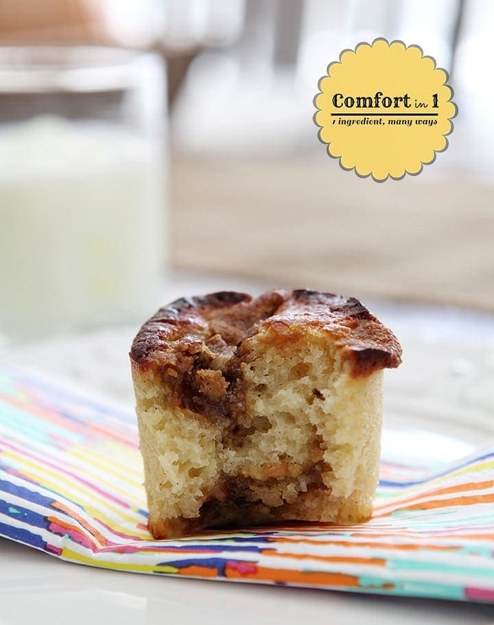 Close up of a halved muffin on a rainbow stripe napkin on a white plate next to a glass of milk on a table
