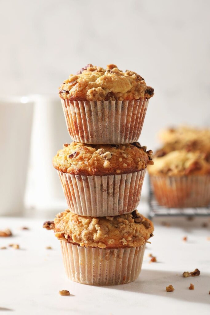 Three Coffee Cake Muffins are stacked on top of each other in front of a coffee mug and a wire cooling rack holding more muffins