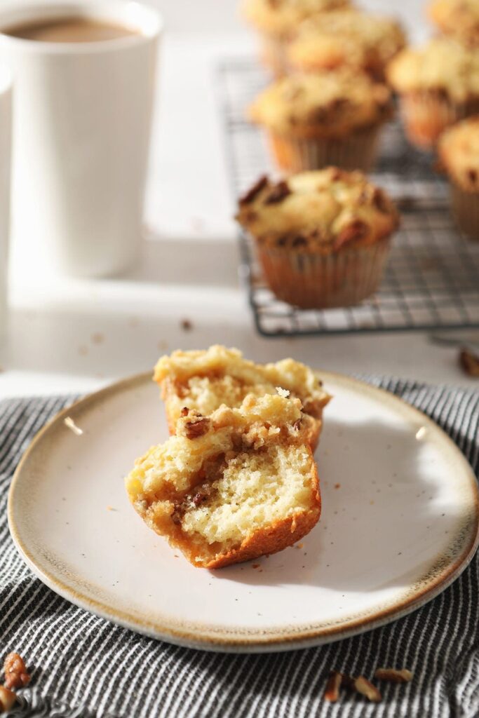 A halved Coffee Cake Muffin sits on a pottery plate in front of coffee mugs and a wire cooling rack holding more muffins