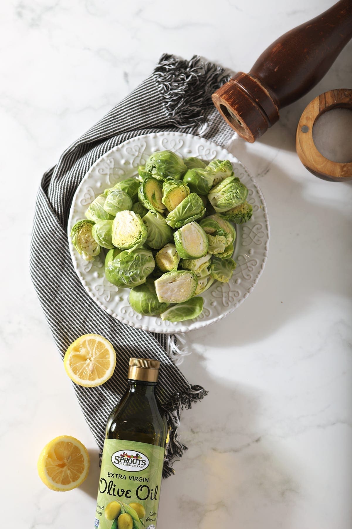 Halved brussels sprouts on a white plate surrounded by olive oil, a lemon, salt and pepper while sitting on a grey striped towel on marble