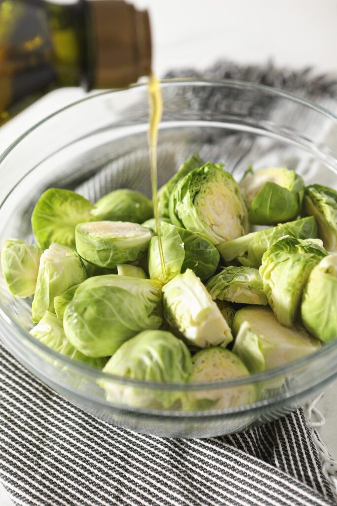 Olive oil drizzles on top of halved brussels sprouts in a glass bowl sitting on a gray striped towel