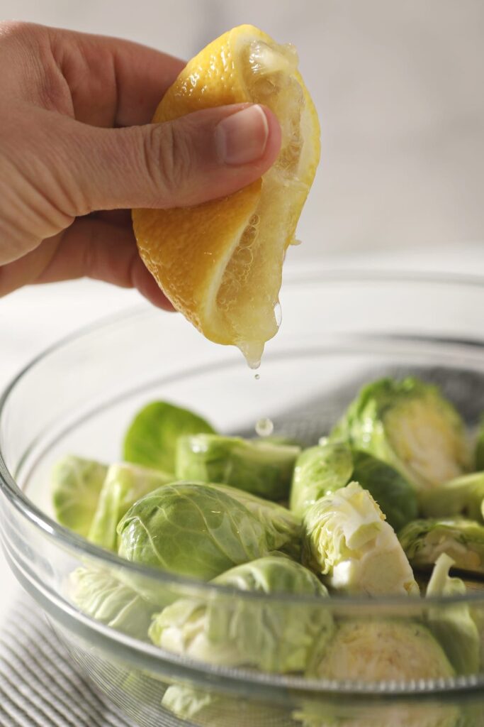 A hand squeezes a lemon on top of halved brussels sprouts in a glass bowl