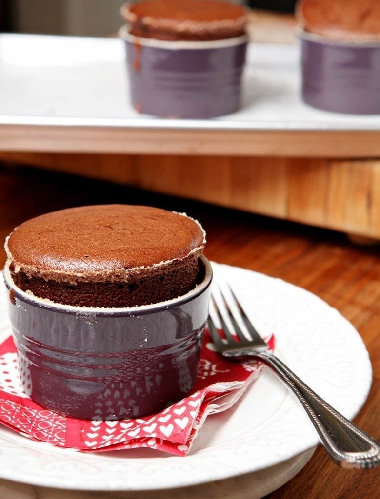 A souffle in a purple dish sits on a red napkin on a white plate in front of other souffles
