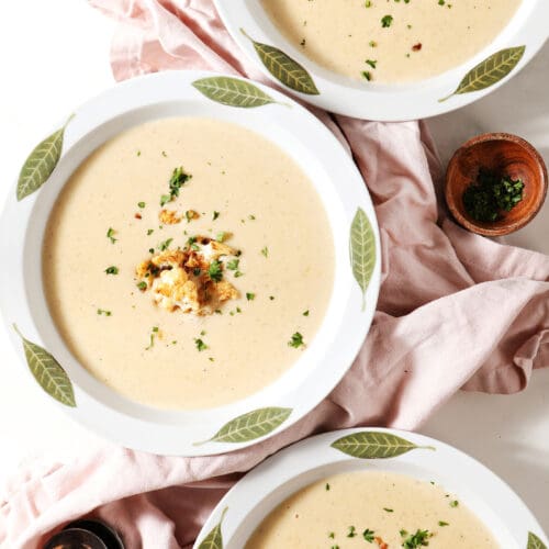 Three white bowls of cauliflower soup garnished with roasted cauliflower and parsley with a pink linen and spoons