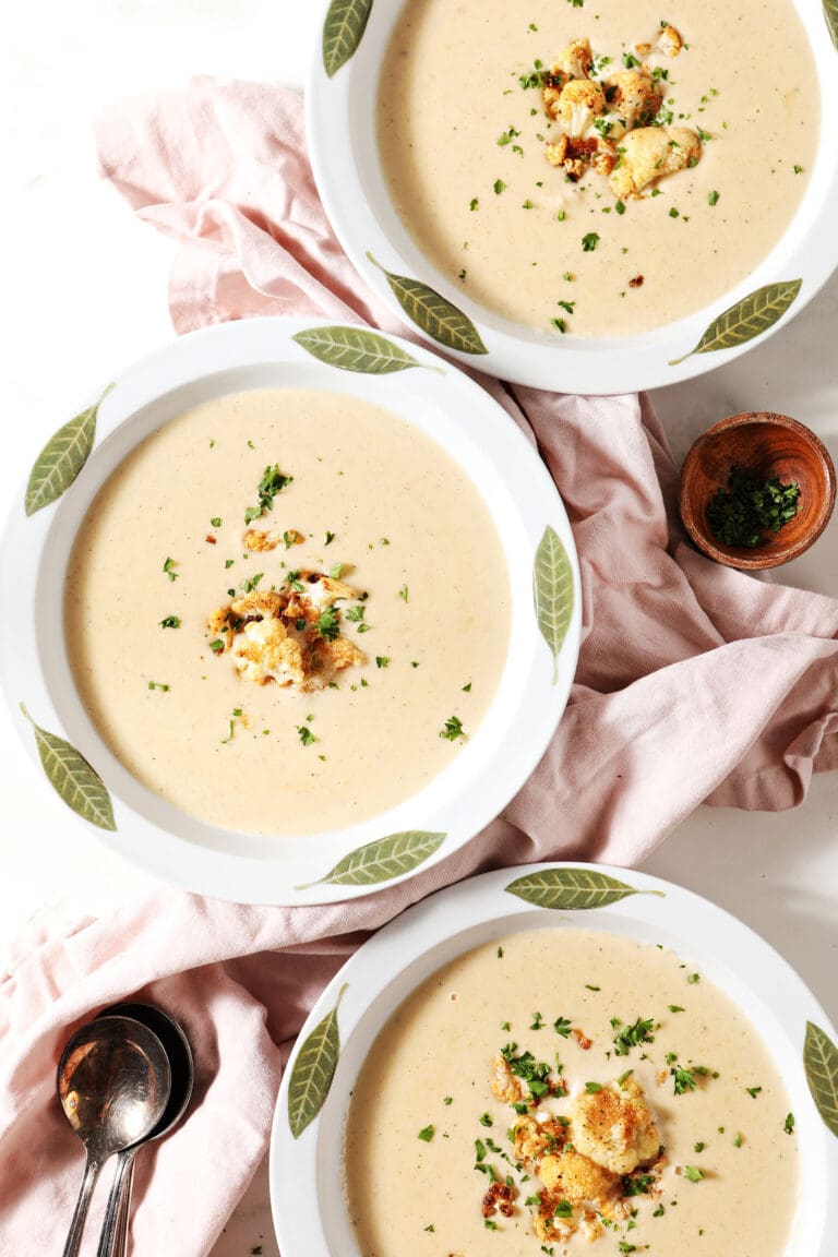 Three white bowls of cauliflower soup garnished with roasted cauliflower and parsley with a pink linen and spoons