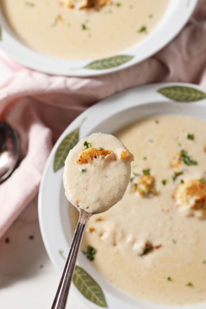 A spoon holds a bite of cauliflower soup above a bowl of it