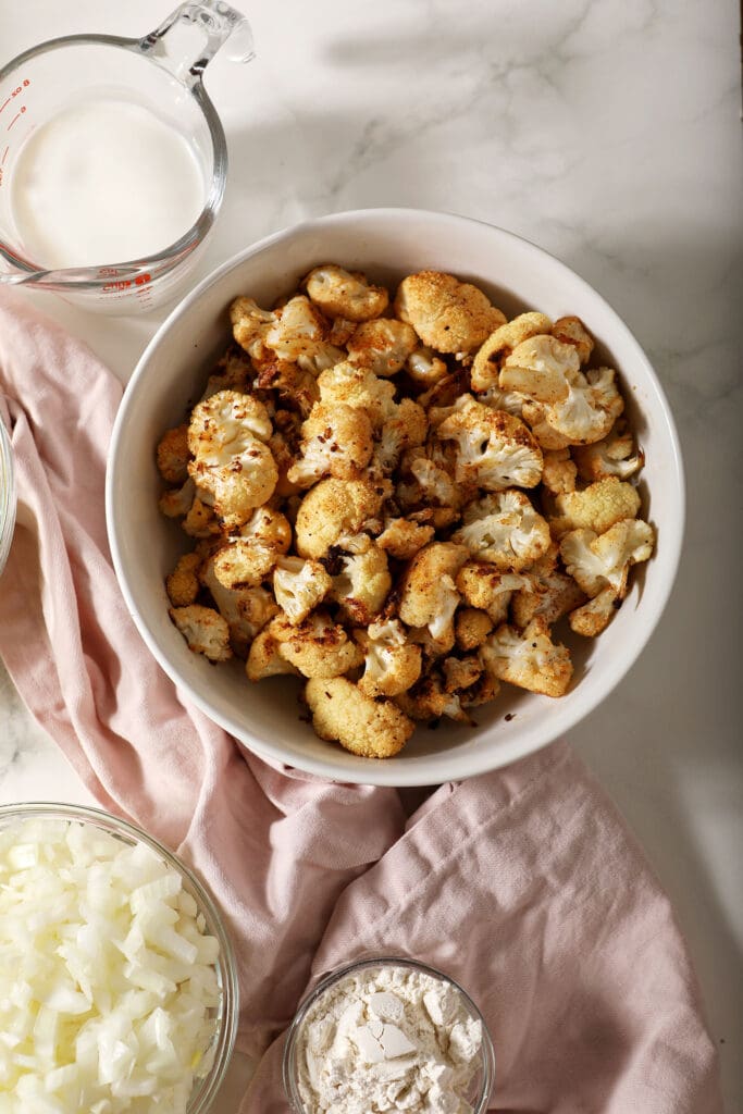 A bowl of roasted cauliflower next to other soup ingredients on marble with a pink linen