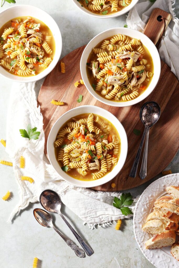 Four bowls of Rotisserie Chicken Noodle Soup on a wooden board with spoons and surrounded by ingredients and sliced bread