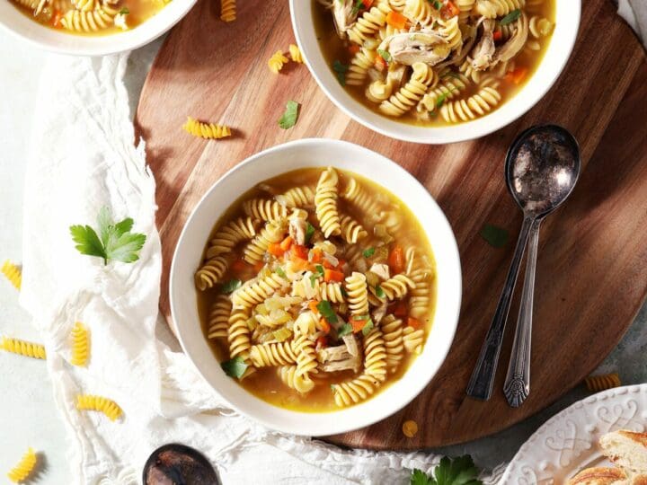 Four bowls of Rotisserie Chicken Noodle Soup on a wooden board with spoons and surrounded by ingredients and sliced bread