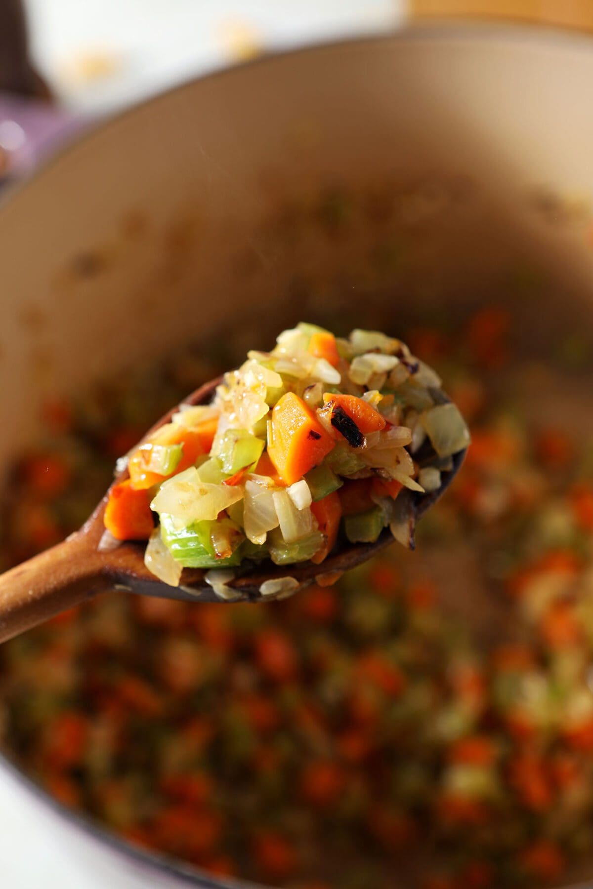 The cooked mirepoix is held up with a wooden spoon over the Dutch oven