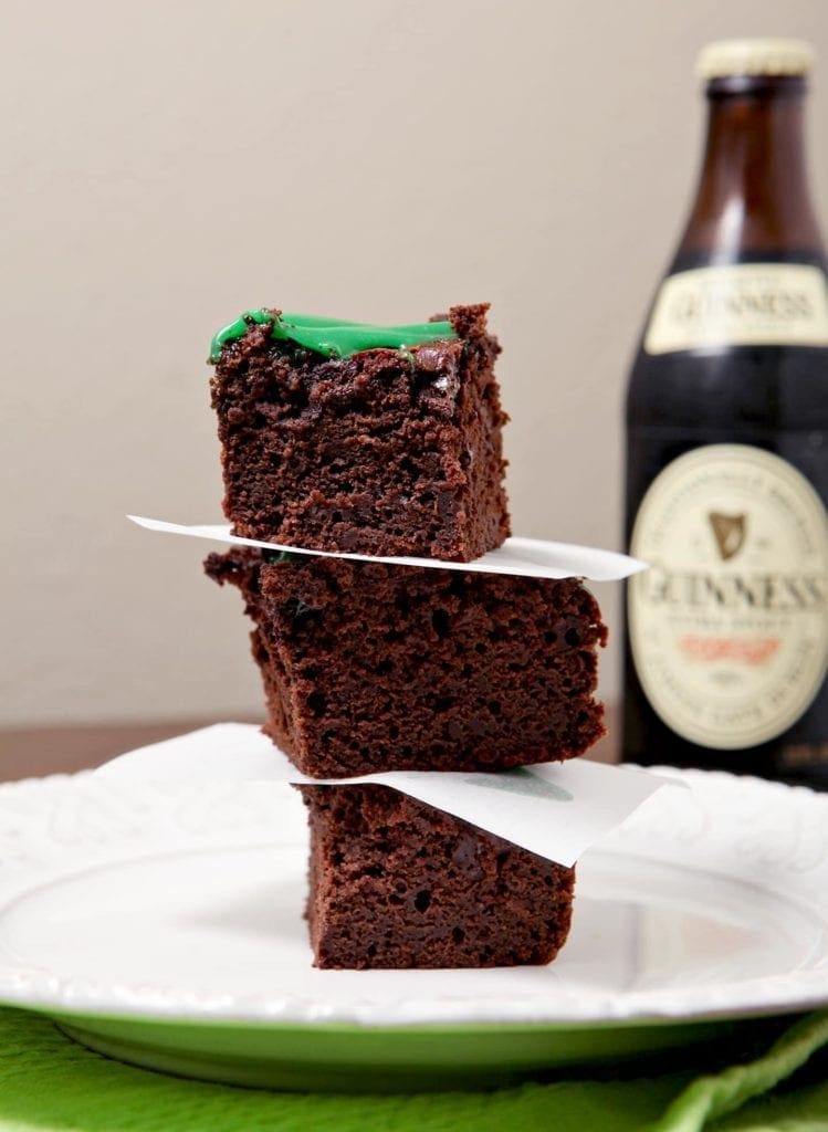 A stack of three brownies sits on a white plate in front of a bottle of Guinness beer