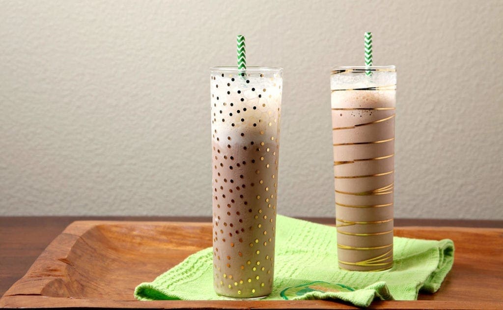 Two gold-decorated glasses holding boozy milkshakes sit on a green towel on a wooden board