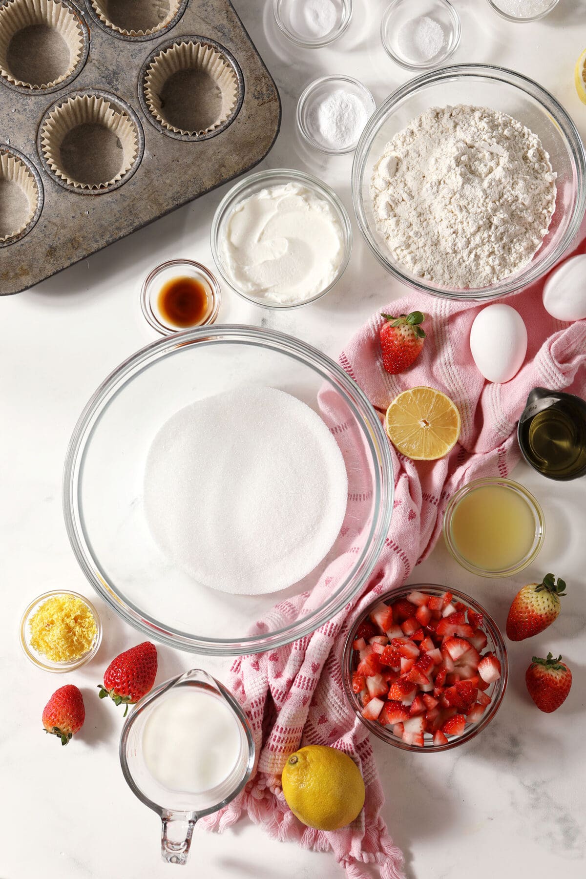 ingredients to make lemon strawberry muffins in bowls on a marble surface with a pink linen and fresh strawberries and lemons
