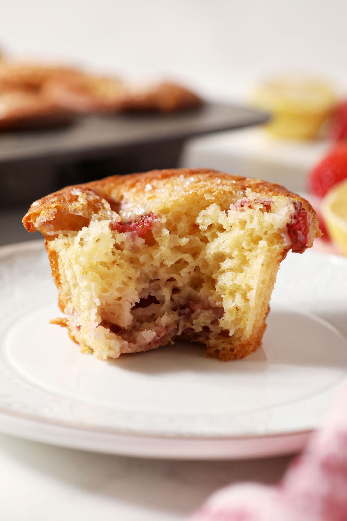 a bitten-into strawberry lemon muffin on a white plate in front of strawberries, lemons and a muffin tin