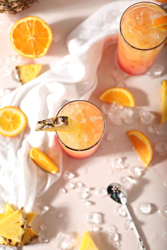 Two glasses holding orange drinks with oranges and pineapples around them