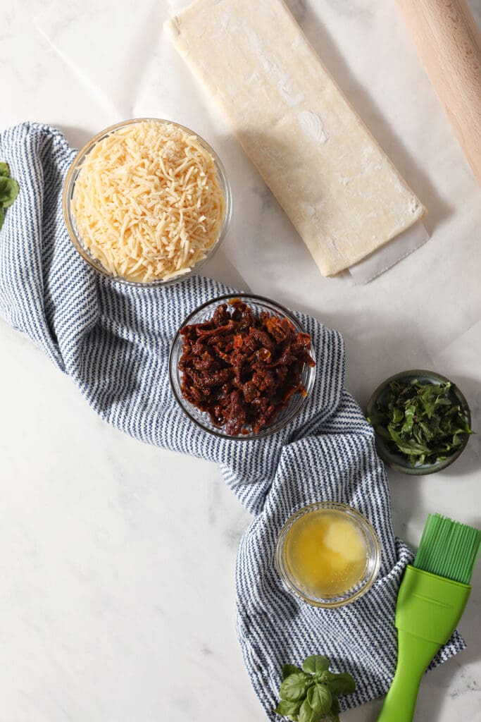 Ingredients to make bruschetta pinwheels in bowls with a blue striped towel on marble