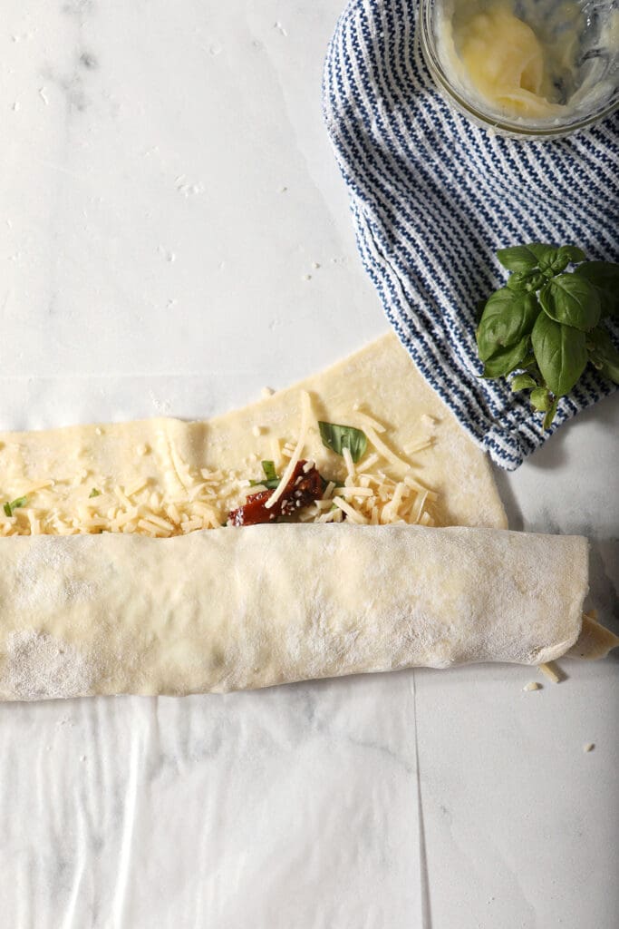 A rolled sheet of puff pastry filled with savory ingredients before sealing and slicing on a marble surface next to a blue striped towel